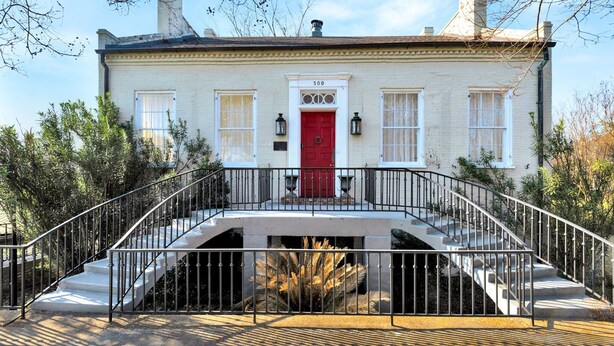 Un edificio de dos pisos con puerta roja y ventanas blancas, una escalera con barandilla metálica y el número "500" sobre la puerta.