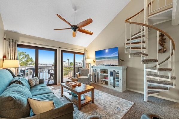 A spacious living room with a large sofa, a coffee table, a ceiling fan, a staircase, and a view of the outdoors. - Cotton Bayou 8C | Waterfront | Boat Slip & Launch | Pool | Beach Getaways (Orange Beach)