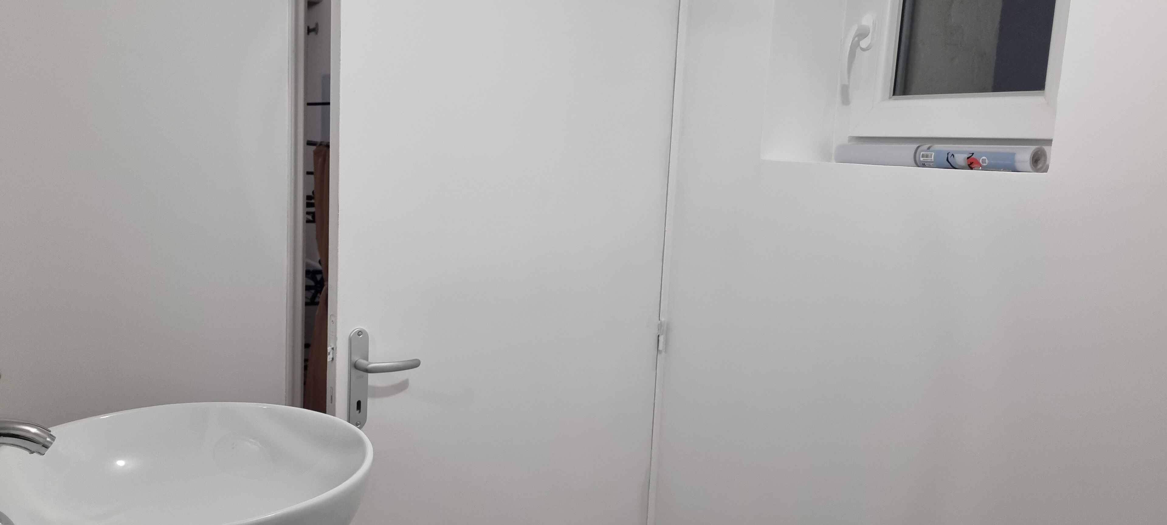 Une salle de bain blanche avec un lavabo, une porte et une petite fenêtre dotée d’un thermomètre.