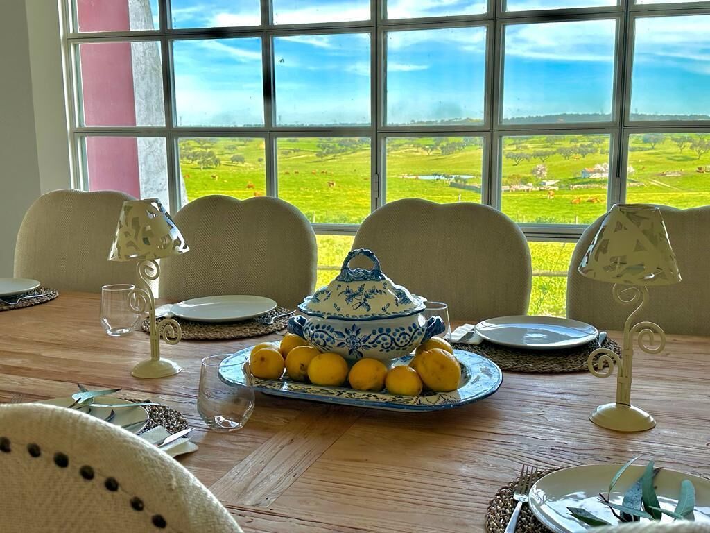 Mesa de comedor lista para cuatro personas, con vista a un paisaje verde a través de un ventanal grande.