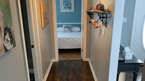 Un estrecho pasillo con suelo de madera, una cama al fondo con pared azul y colcha blanca, y un pequeño estante con objetos decorativos.