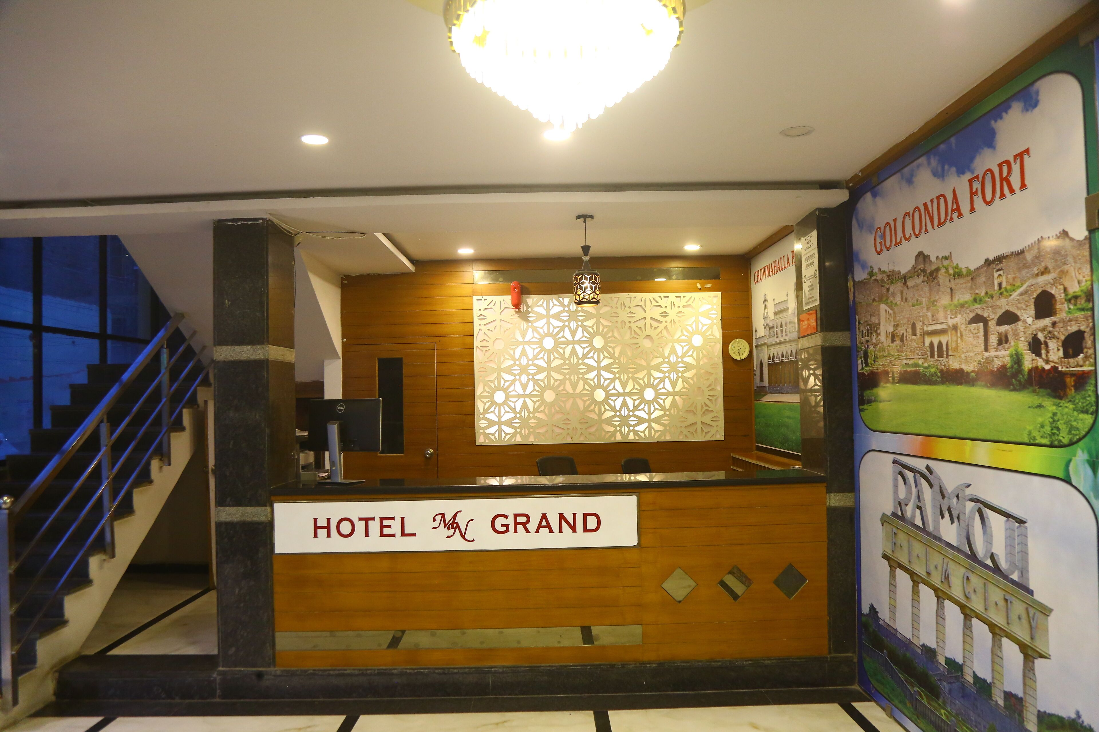 Zona de recepción de un hotel con un mostrador de madera que dice "HOTEL GRAND", un candelabro decorativo y una pared con un cartel del Fuerte de Golconda.