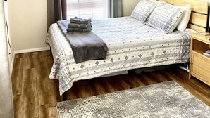 Un dormitorio ordenado con cama, una mesita de noche, una ventana con cortinas, un cuadro en la pared y una alfombra estampada.
