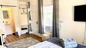 Un dormitorio con cama, mesita de noche, televisor, ventana con cortinas, armario y baño con lavamanos y espejo.