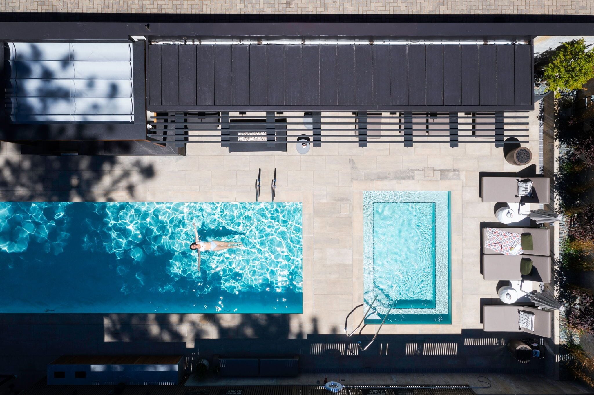 Vista aérea de una zona de piscina en la azotea, que incluye una piscina rectangular grande, una piscina cuadrada más pequeña y un área de descanso con sillones y sombrillas.