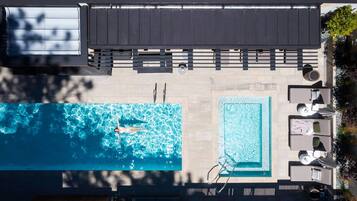 Vista aérea de una zona de piscina en la azotea, que incluye una piscina rectangular grande, una piscina cuadrada más pequeña y un área de descanso con sillones y sombrillas.