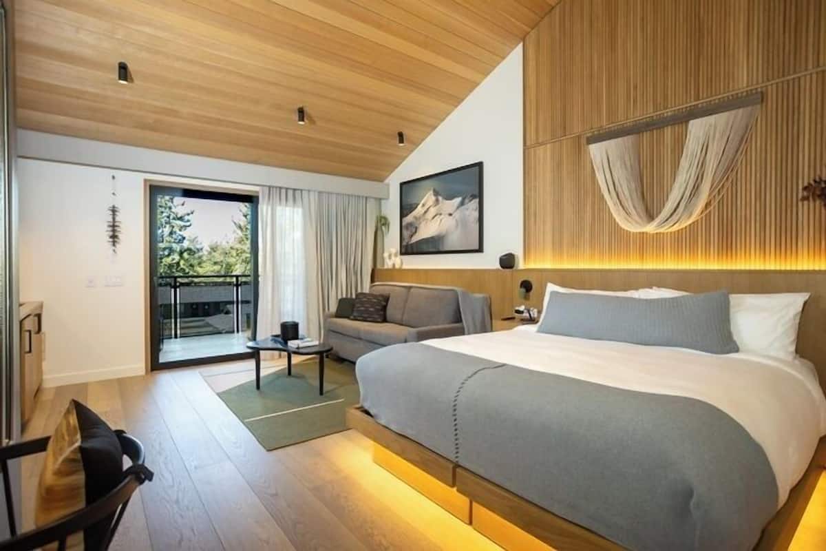 A modern bedroom with a large bed, a sofa, a small table, and a sliding glass door leading to a balcony.