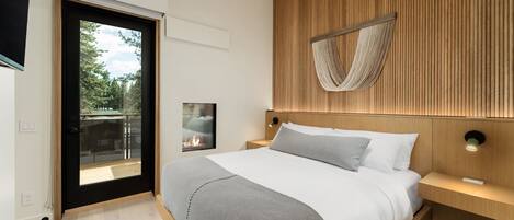 A modern bedroom with a large bed, a wooden headboard, a small bedside table with a lamp, and a view of the outdoors through a sliding door.