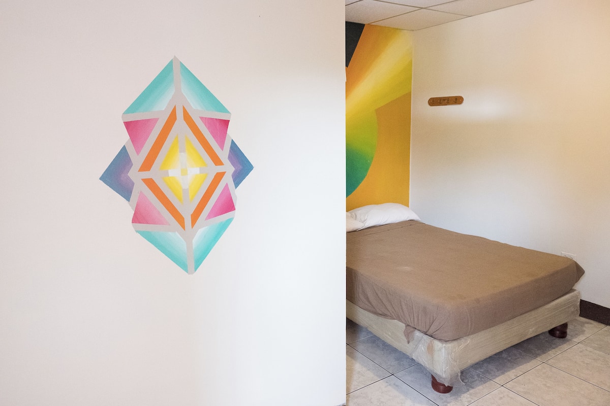 A room with a bed, a colorful wall decoration, and a small wooden sign on the wall.