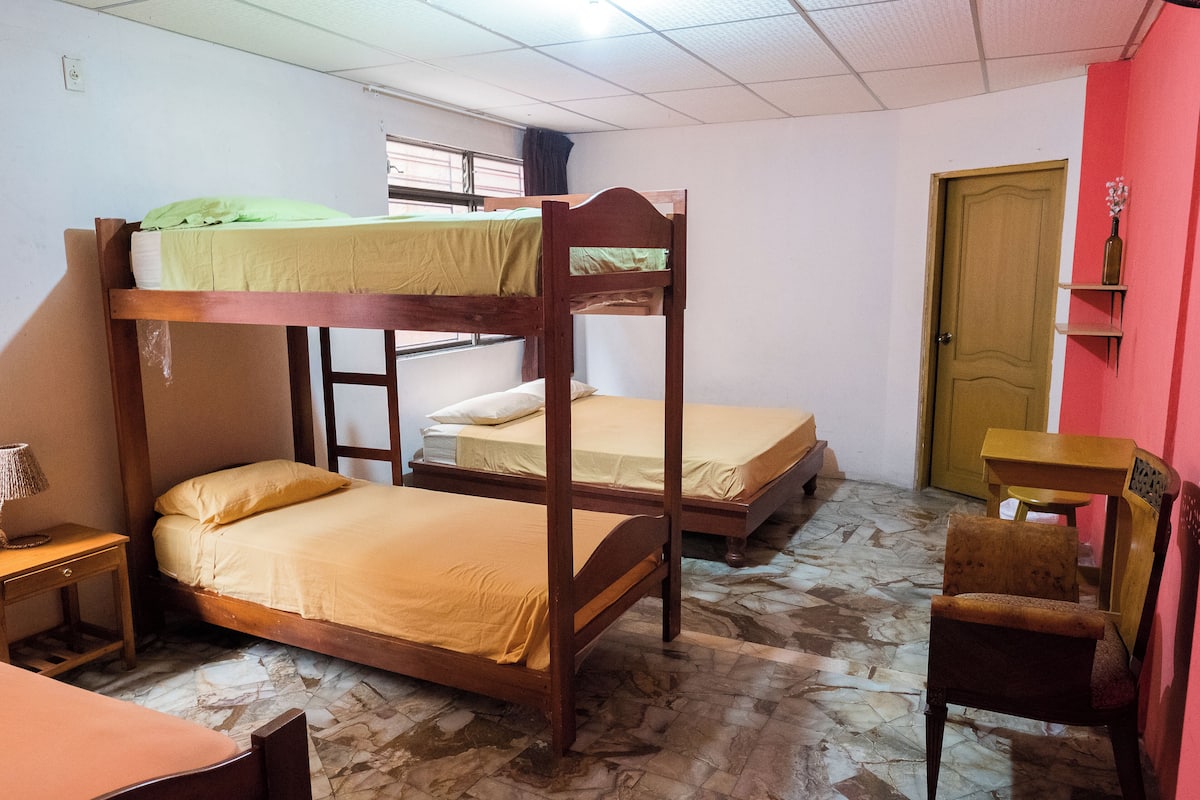 A dormitory room with bunk beds, a wooden desk, a chair, and a small table.