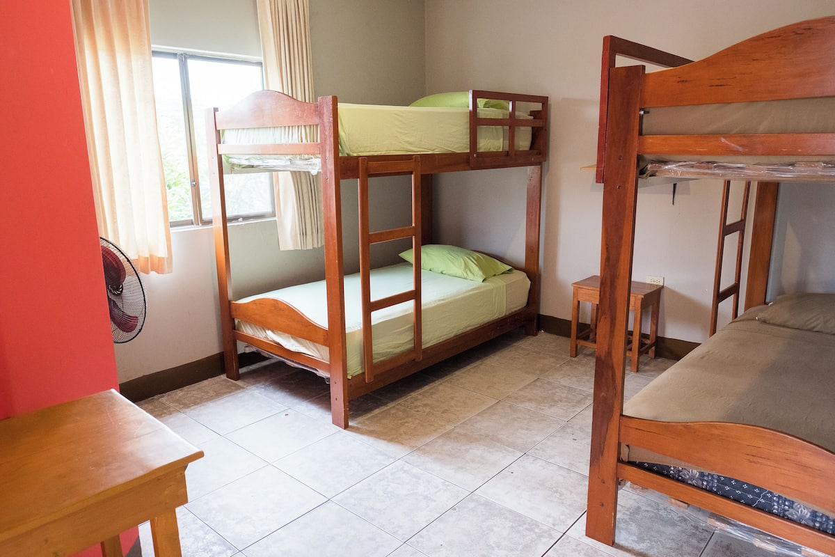 A room with two bunk beds, a small wooden table, a standing fan, and a window with curtains.