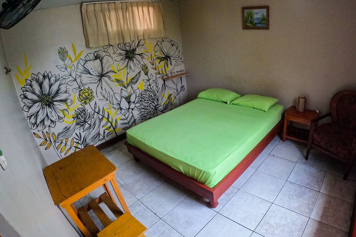 A bedroom with a bed, a chair, a small table, a wall with a floral mural, a window with curtains, and a picture on the wall.
