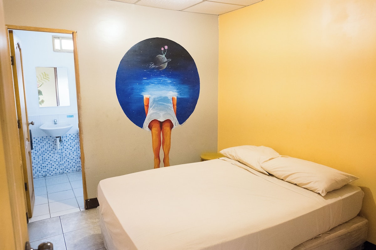 A bedroom with a bed, a wall mural, and a bathroom visible through an open door.