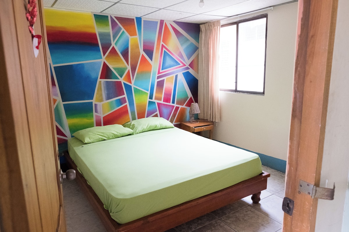 A bedroom with a colorful geometric wall design, a green bed, a wooden nightstand, and a window with curtains.