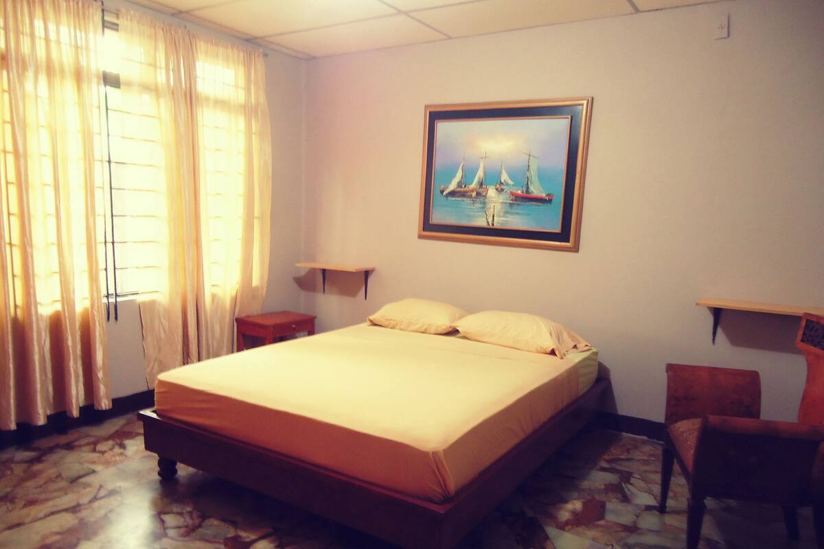 A hotel room with a bed, a chair, a nightstand, a painting of boats on the wall, and a window with curtains.