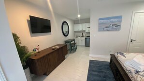 Un dormitorio moderno con cama, una consola de madera, televisión y una cocina al fondo.