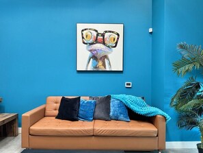 A room with a blue wall, a tan sofa with multiple cushions, a framed artwork above the sofa, and a potted plant to the right. - Spacious 2BR ,Family& Pet Friendly, Tesla charging Station on site, Near Disney (Santa Ana)