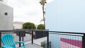 Un balcon agrémenté d’un fauteuil Adirondack turquoise, d’une rambarde en métal noir et offrant une vue sur un immeuble au toit rouge.