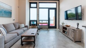 Un salon moderne avec un canapé, une table basse et un téléviseur. De grandes fenêtres laissent entrer la lumière naturelle et offrent une vue sur l’extérieur.