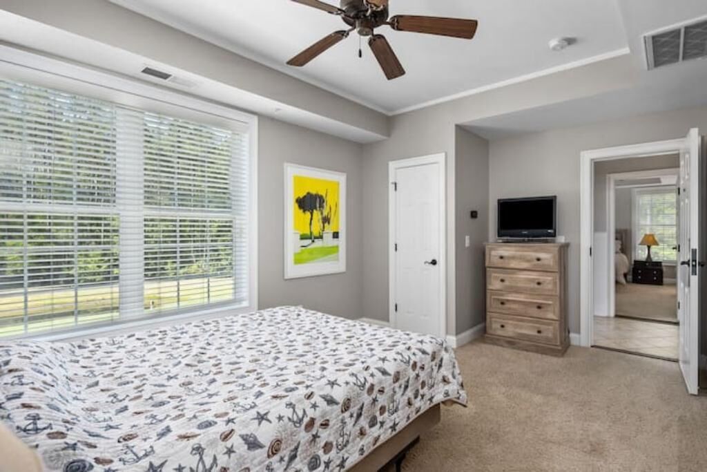 Un dormitorio con cama, ventilador de techo, televisor, cómoda y puerta que da a otra habitación.