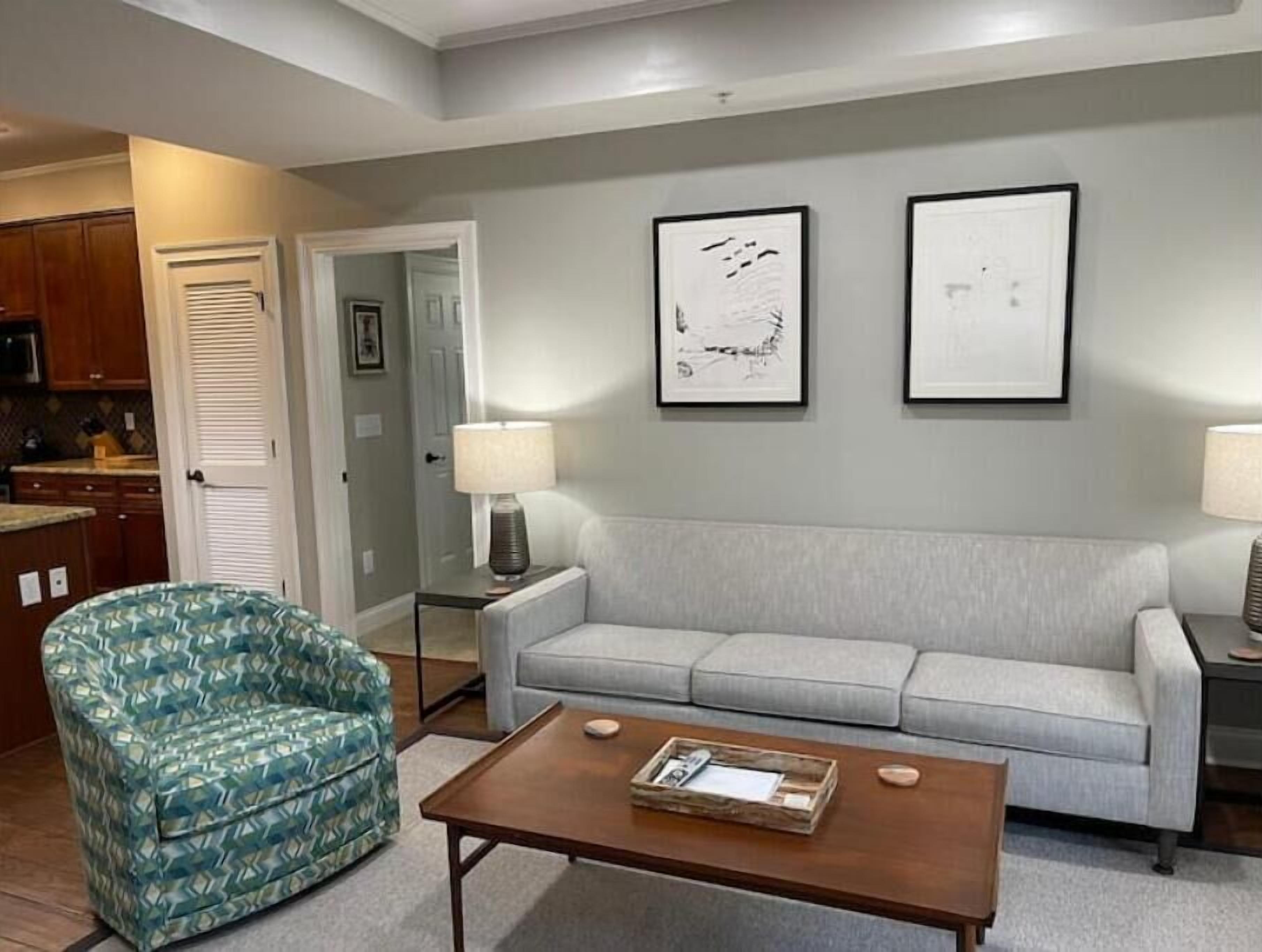 A living room with a patterned armchair, a grey sofa, a coffee table, and two framed artworks on the wall.