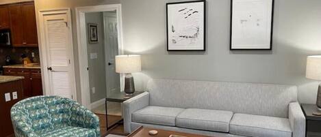 A living room with a patterned armchair, a grey sofa, a coffee table, and two framed artworks on the wall.