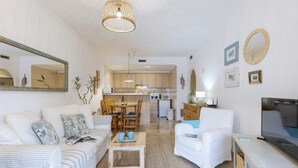 A modern living room with a sofa, armchairs, a coffee table, and a TV. A kitchen is visible in the background with wooden cabinets and a dining area. - Olive Beach (Javea / Xàbia)