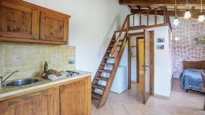 Una cocina con armarios de madera, salpicadero de azulejos, un lavabo y una estufa. Una escalera de madera conduce a un dormitorio con una cama y una mesita de noche.