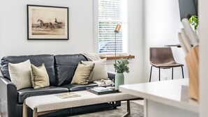A modern living room with a black leather sofa, a coffee table, a framed picture, and a window with blinds. - Sage at The Shire 255 - Unit A - Downtown! (Southern Pines)