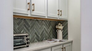 A kitchen with white cabinets, a herringbone-patterned backsplash, a white vase with flowers, and a toaster on the counter. - Sage at The Shire 255 - Unit A - Downtown! (Southern Pines)