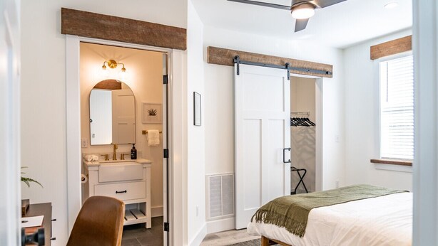 A bedroom with a bed, a desk, a chair, a wardrobe, and a bathroom with a mirror and sink. - Sage at The Shire 255 - Unit A - Downtown! (Southern Pines)