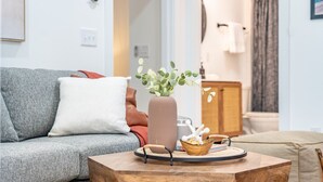 Een woonkamer met een grijze bank, een houten salontafel met een vaas en decoratieve objecten, en een uitzicht op een badkamer op de achtergrond.