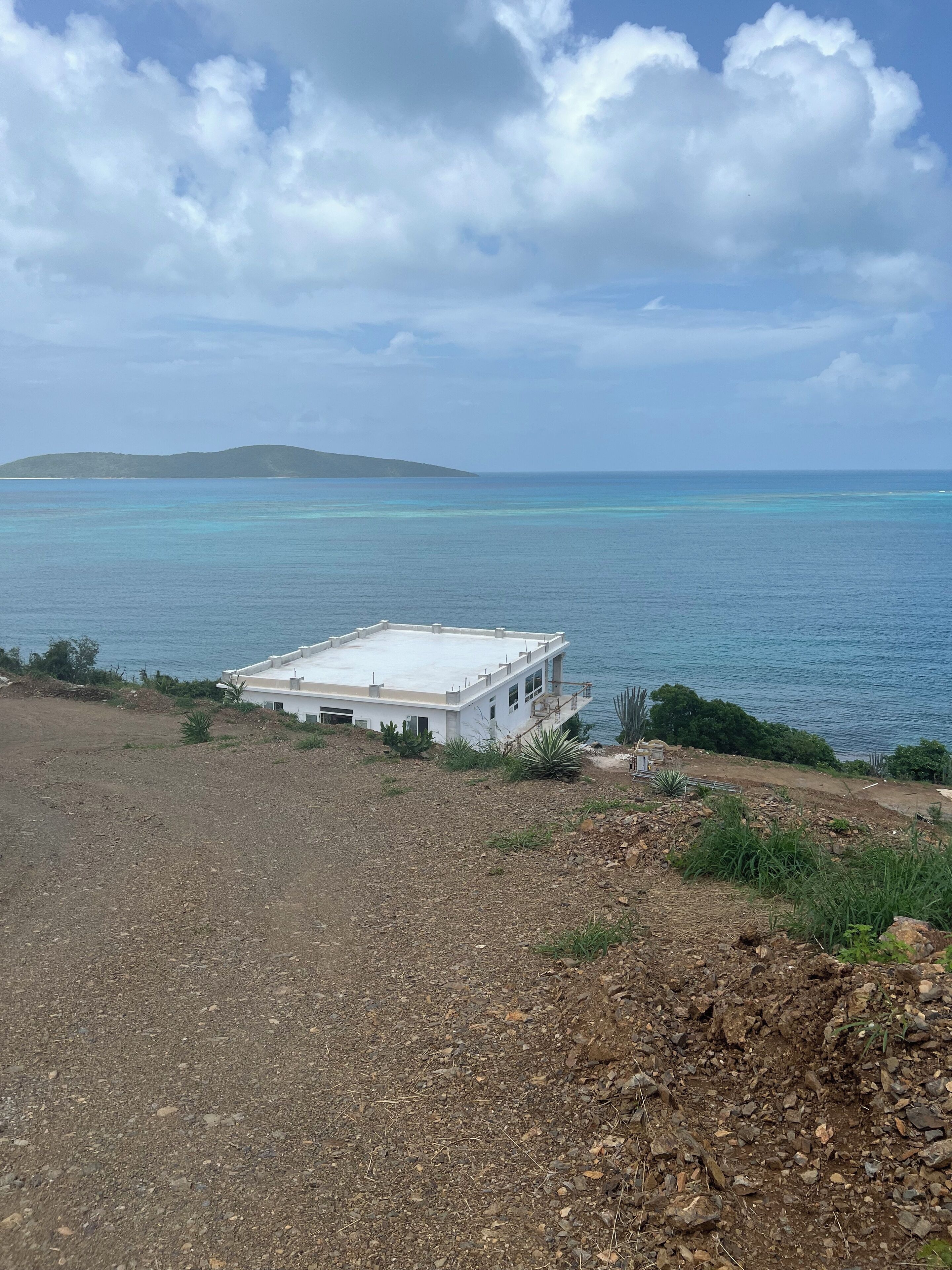 A white building with a flat roof near a body of water, surrounded by rocky terrain and sparse vegetation.