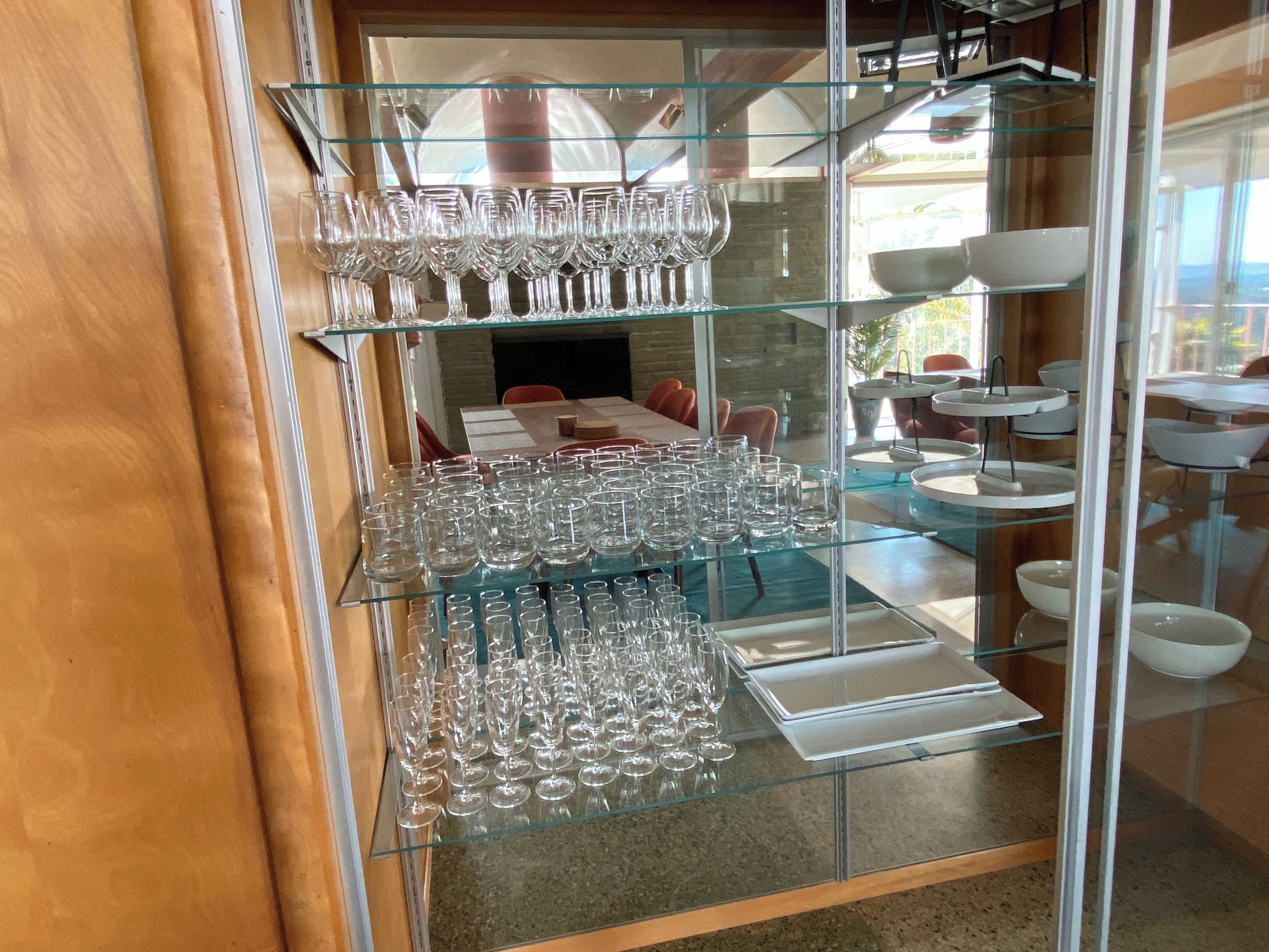 A glass display case with multiple tiers of wine glasses and plates, set against a backdrop of a dining area with tables and chairs.