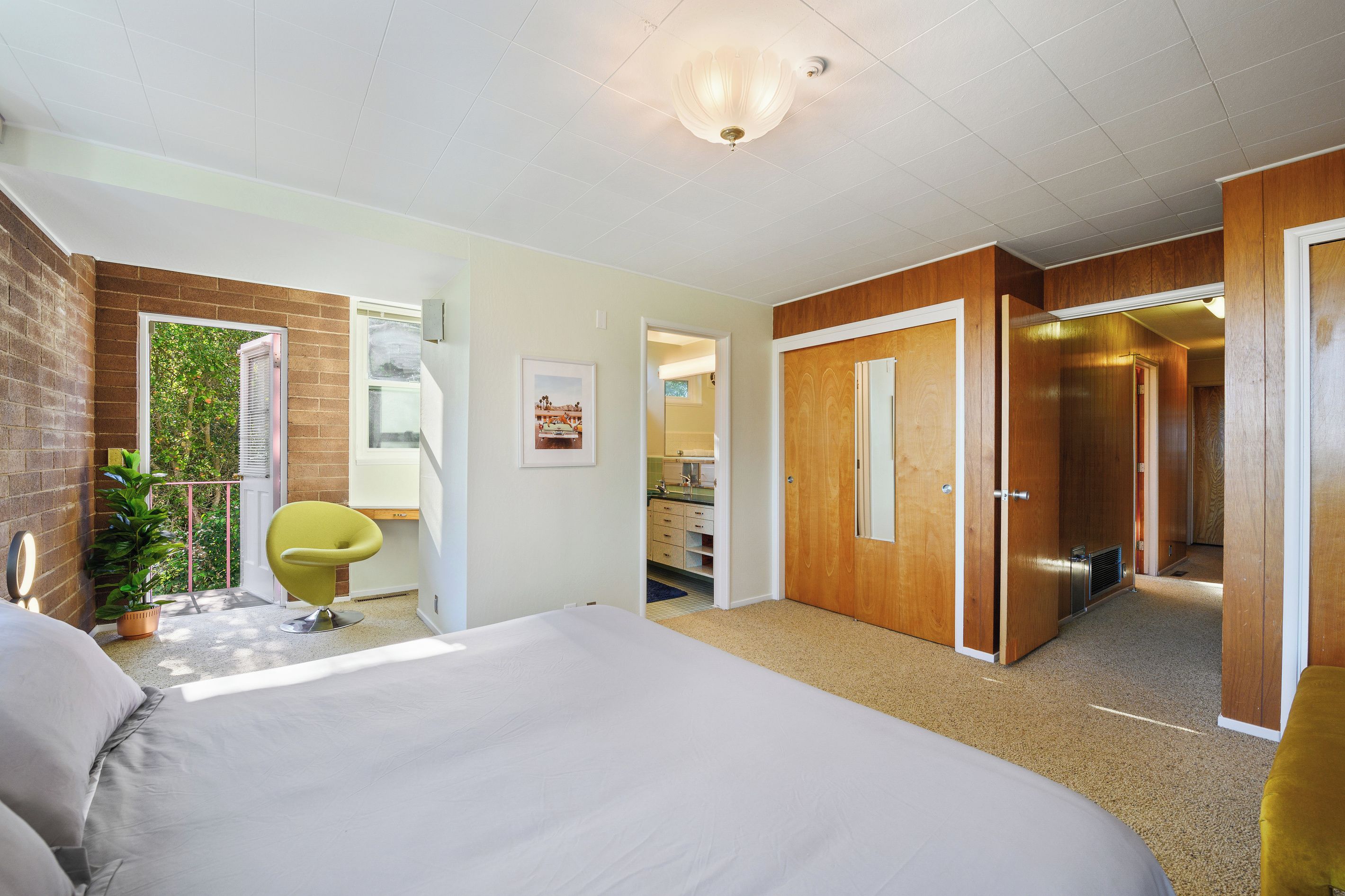 A bedroom with a bed, a chair, a potted plant, a door, and a view of a bathroom.