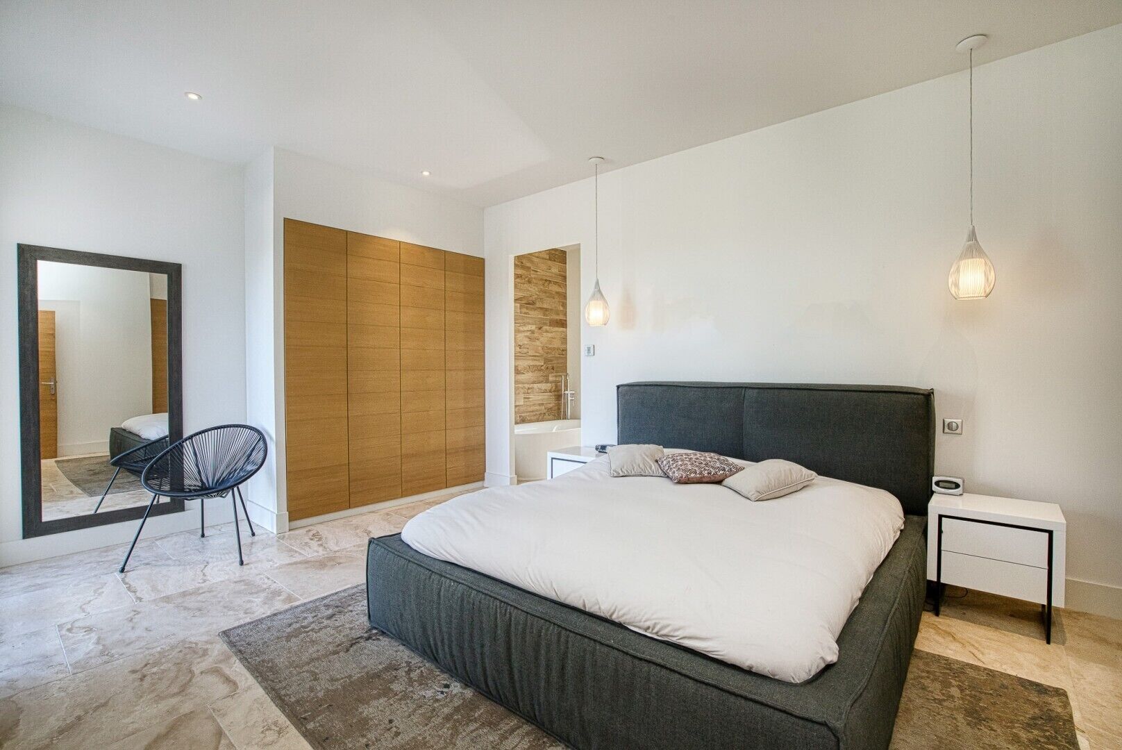 A modern bedroom with a large bed, a chair, a wardrobe, and a mirror.