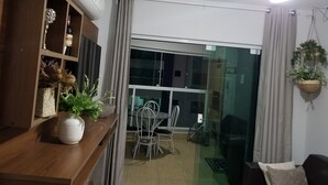 Un salon moderne avec une étagère en bois, une table à manger avec des chaises, une porte coulissante en verre donnant sur un balcon et un ventilateur de plafond.