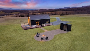 Una cabaña moderna con terraza, un brasero y un ventanal grande con vista a un campo cubierto de césped.