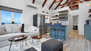 Ein modernes Wohnzimmer mit Sofa, Couchtisch und Hocker, eine Küche mit Barhocker und Blick ins Freie.
