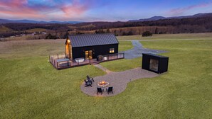 Eine moderne Hütte mit einer Terrasse, einem Feuerkorb und einem großen Fenster mit Blick auf eine Wiese.