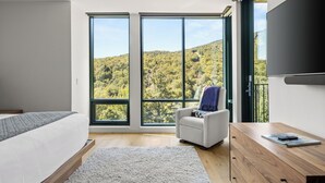 A bedroom with a large window, a bed, a chair, a wooden dresser, and a TV.