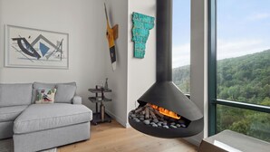 A modern living room with a grey sofa, a fireplace with logs, a framed artwork, and a view of a forest through a large window.
