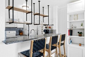 A modern kitchen with a granite countertop, black chairs with woven seats, and a white shelving unit with decorative items.