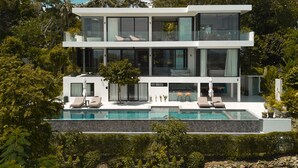 Une maison moderne à plusieurs étages, dotée d’une piscine, entourée d’une végétation luxuriante et de palmiers.