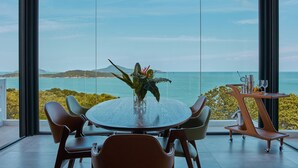 Un espace repas moderne avec une table ronde et des chaises, un vase de fleurs et une vue sur la mer grâce à de grandes fenêtres.