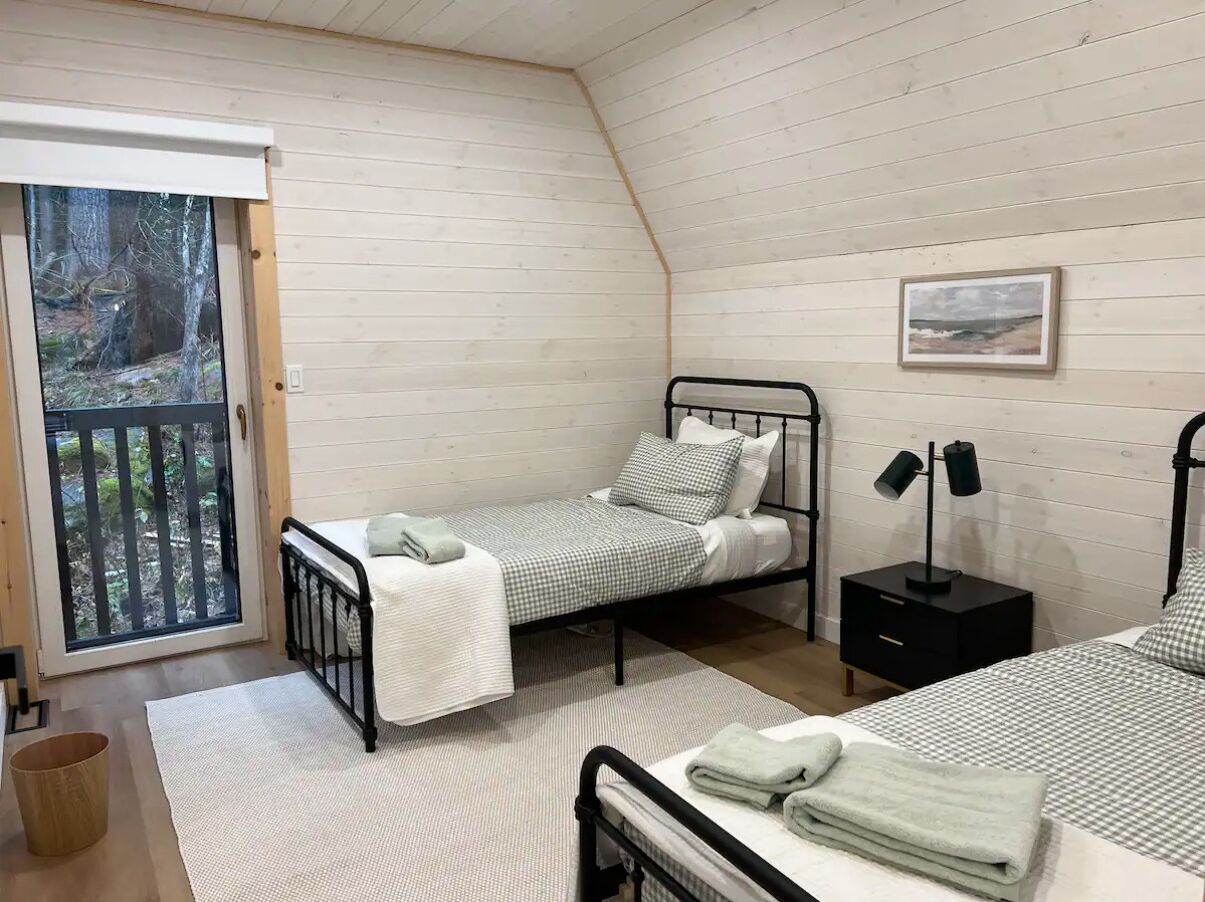 Ein Schlafzimmer mit zwei Einzelbetten, einem kleinen schwarzen Nachttisch mit Lampe, einem Fenster mit Blick auf Bäume und einer Holzbalkendecke.