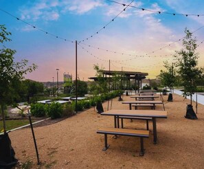 An outdoor seating area with picnic tables and string lights, surrounded by greenery and a view of a building in the background.