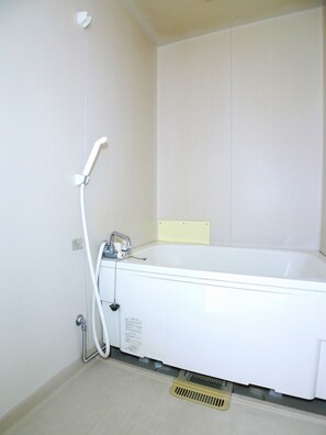 A white bathtub with a showerhead and hose, a yellow sign on the wall, and a vent on the floor. - Silk Village (Itako)