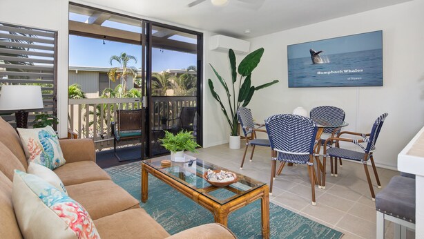 A spacious living room with a sofa, coffee table, and dining area with chairs. There is a large window with a view of palm trees and a balcony.