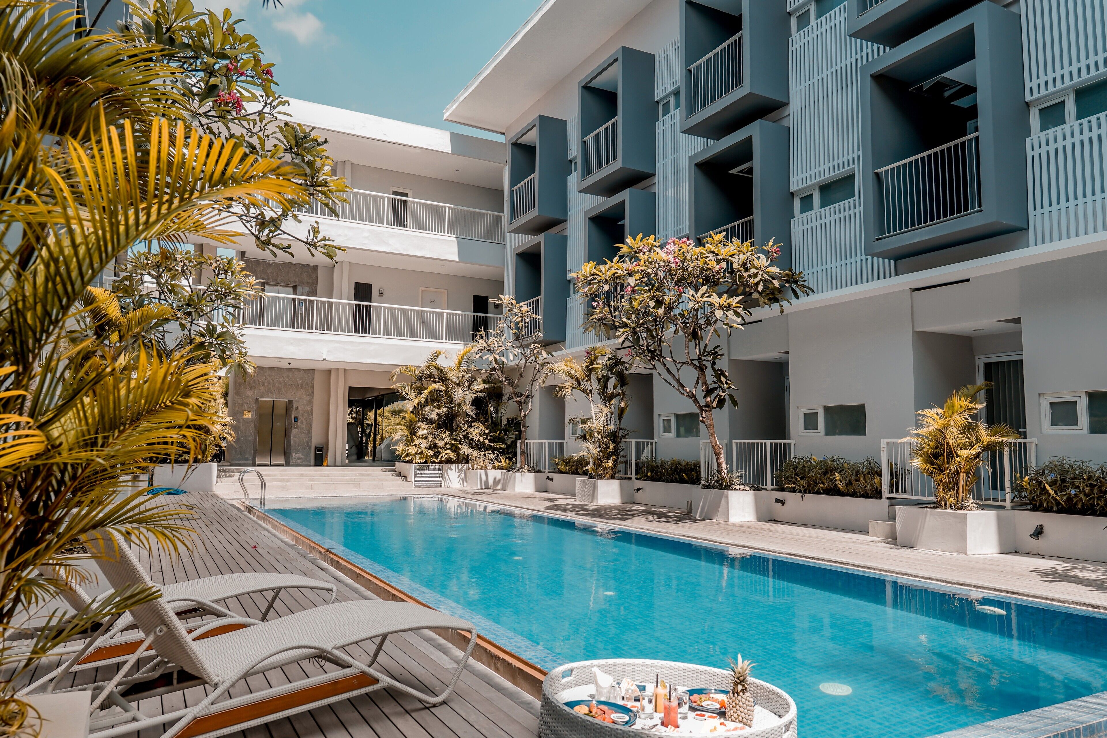 Une piscine entourée de chaises longues, elle-même bordée d’un immeuble à plusieurs étages doté de balcons et agrémenté de plantes en pot.